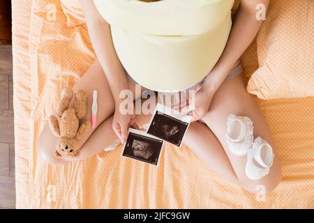 Donna incinta seduta a letto e guardando Ultrasound scansione foto del suo bambino. Una donna incinta mostra il suo rapporto ecografico e il suo feto. Foto Stock