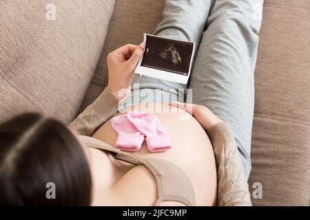 Donna incinta seduta a letto e guardando Ultrasound scansione foto del suo bambino. Una donna incinta mostra il suo rapporto ecografico e il suo feto. Foto Stock