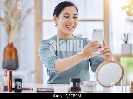 Ragazza allegra seduta alla sua scrivania prendendo un selfie sul suo cellulare prima di applicare prodotti per la cura della pelle. Sorridente donna che usa il suo smartphone a. Foto Stock