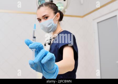 donna piuttosto giovane in un vestito chirurgico in guanti di gomma blu sullo sfondo di una lampada chirurgica tiene una siringa nelle sue mani. Foto Stock