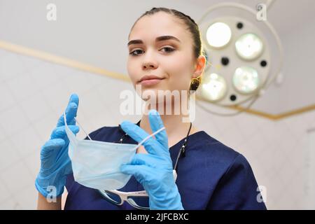 bella giovane donna in un abito chirurgico in guanti di gomma blu sullo sfondo di una lampada chirurgica mette su una maschera sterile. Foto Stock