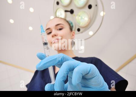 piuttosto giovane medico donna in un vestito chirurgico in guanti di gomma blu sullo sfondo di una lampada chirurgica tiene una siringa nelle sue mani primo piano. Foto Stock