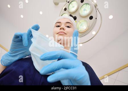 bella giovane donna in un abito chirurgico in guanti di gomma blu sullo sfondo di una lampada chirurgica mette su una maschera sterile. Foto Stock