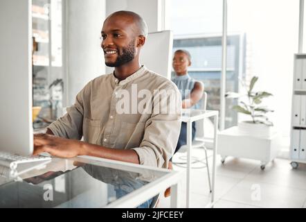 Giovane uomo d'affari afro-americano felice che usa un computer desktop in un ufficio al lavoro. Soddisfare uomini d'affari digitando un email ad una scrivania Foto Stock