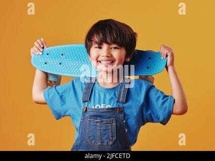 Un adorabile ragazzo asiatico che guarda felice mentre tiene il suo skateboard su uno sfondo arancione. Ragazzo carino che indossa abiti casual sorridendo come lui Foto Stock