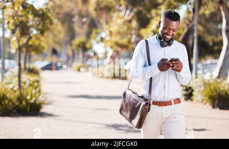 Un uomo afro-americano che indossa cuffie e SMS su uno smartphone mentre si è in città per spostarsi ogni giorno. Maschio nero sorridente durante il controllo Foto Stock