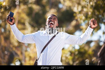L'uomo afro-americano festeggia una vittoria mentre usa il suo cellulare e indossa le cuffie. Felice giovane maschio che esprime successo e vittoria mentre cammina Foto Stock