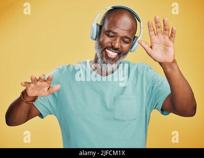 Felice uomo africano maturo ballare mentre si ascolta la musica con le cuffie su uno sfondo giallo studio. Spensierato uomo nero senior con grigio Foto Stock