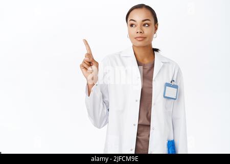 Concetto di assistenza sanitaria. Sorridente giovane terapeuta femminile, chirurgo in cappotto bianco, puntando e guardando a sinistra copyspace, in piedi su sfondo bianco Foto Stock