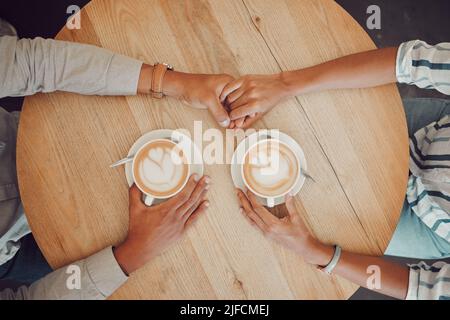 Primo piano di una giovane coppia innamorata che tiene le mani con due tazze di caffè su un tavolo mentre si siede in un bar. Mani maschili e femminili con cappuccino caldo Foto Stock