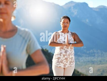 Donna matura meditando con mani unite e occhi chiusi respirando profondamente. Persone mature che fanno yoga insieme in natura in una giornata di sole Foto Stock