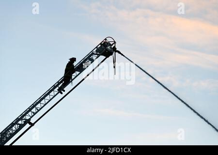 Seattle - 30 giugno 2022; vigile del fuoco su un camion a scala che spruzza il flusso d'acqua in silhouette contro un cielo di moro fumé Foto Stock