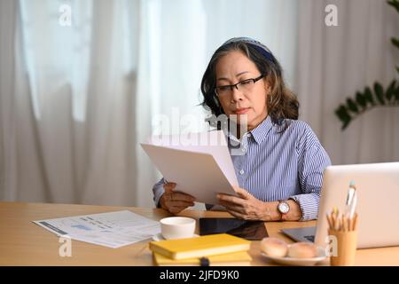 Donna asiatica mature focalizzata indossando occhiali utilizzando il laptop e la gestione della finanza domestica Foto Stock