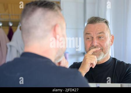 Uomo anziano che guarda il suo riflesso in uno specchio del bagno con un sorriso rueful in una vista sopra la spalla Foto Stock