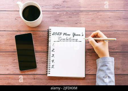 Mani che scrivono una lista di risoluzioni di Capodanno 2023 con tazza di caffè e smartphone Foto Stock