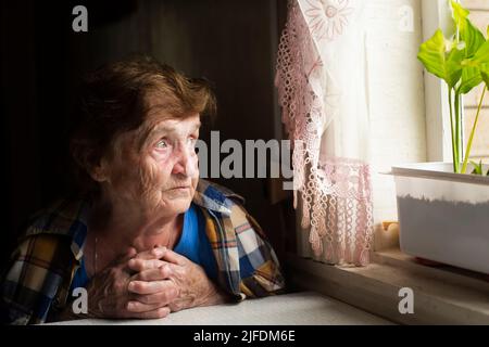 Una vecchia donna solitaria fissa doomfully fuori la finestra. Foto Stock