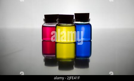 Tre vasetti di vetro con tappi in sughero con i tre colori primari, magenta, blu e giallo in una retroilluminazione che si riflettono su una superficie a specchio Foto Stock