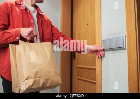 Uomo di consegna in maschera protettiva che tiene il sacchetto di carta con il cibo nell'ingresso. Il corriere si trova alla porta e suona il campanello del cliente. Lui Foto Stock