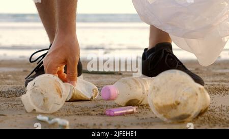 Primo piano del volontario maschile raccogliere e raccogliere rifiuti sulla spiaggia. L'uomo pulisce la costa dai rifiuti di plastica - bottiglie, tazze. Volontari Foto Stock