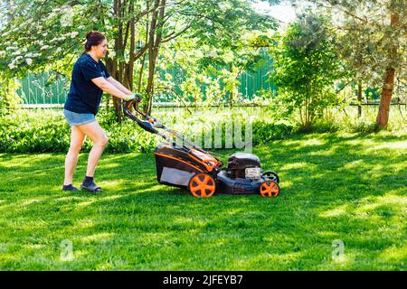 Giardiniere femmina dai capelli castani in abiti casual con rasaerba a benzina o tagliaerba in giardino. Attività nel fine settimana Foto Stock