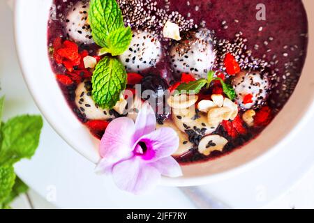 Deliziosa ciotola di frullato con frutta esotica tropicale, bacche, semi, decorata con orchidee e menta verde. Il concetto di nutrizione sana. Superficie piana. Foto Stock