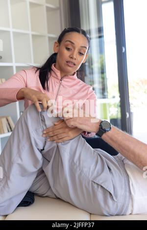 Fisioterapista femminile biracale che dà la terapia di massaggio delle gambe all'anziano caucasico a casa Foto Stock