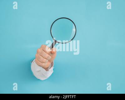 Una mano della donna che sporge da un foro in uno sfondo blu di carta tiene una lente d'ingrandimento. Foto Stock