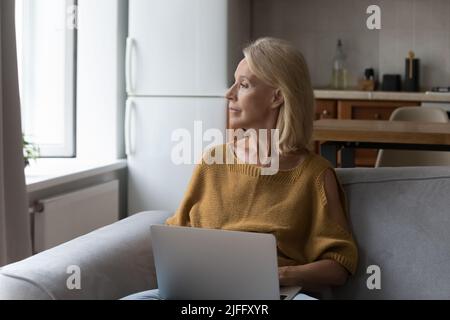 Donna anziana e pensiva seduta sul divano con un computer portatile Foto Stock