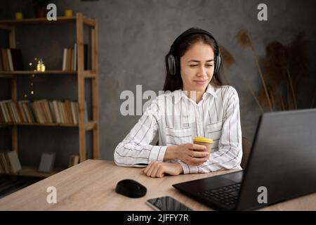 Donna ispanica executive che ha una videochiamata con un laptop e cuffie in un ufficio domestico, ragazza che guarda video conferenza e bere caffè Foto Stock