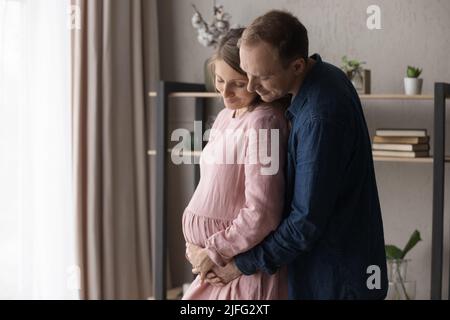 Amare i genitori futuri che aspettano il loro primo bambino che abbraccia in piedi all'interno Foto Stock