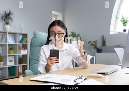 Giovane bella donna d'affari asiatica che lavora in ufficio, sorridente e felice di vittoria, usando il telefono, leggendo buone notizie, celebrando trionfo. Foto Stock