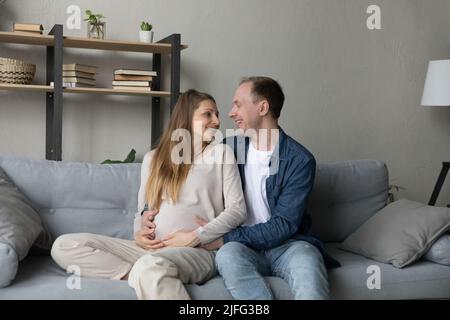 La moglie incinta guarda il marito con amore seduto sul divano Foto Stock