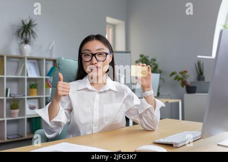 Bella donna di affari sorridente e guardando la macchina fotografica, donna asiatica che offre la carta di banca ai clienti, mostrando i pollici in su con la mano, donna d'affari che lavora in ufficio. Foto Stock
