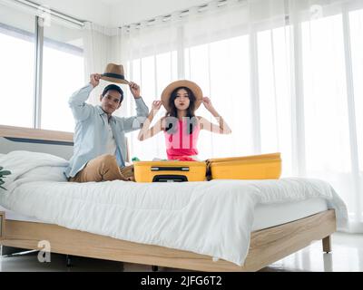 Coppia di viaggiatori che si preparano a partire. Giovane donna asiatica e uomo in denim camicia con cappelli da spiaggia mentre si impacchettano con valigie gialle sul letto nel letto Foto Stock