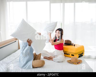 Coppia cuscino lotta. Asian bella giovane donna e uomo in denim camicia seduta e lotta con cuscini vicino alla valigia gialla sul letto Foto Stock