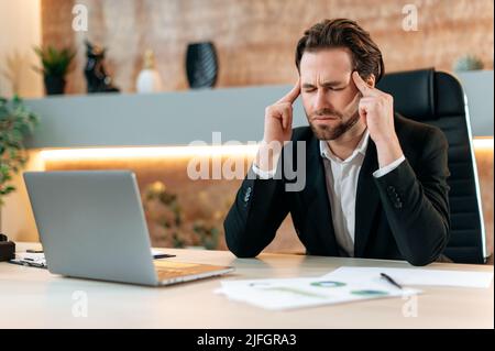 Affaticamento l'uomo d'affari caucasico, impiegato di ufficio, siede ad un posto di lavoro in un ufficio moderno, tocca i suoi templi, testa di massaggio, tatto sollecitato, overworked, mal di testa, emicrania, bisogno di riposo e caffè brak Foto Stock