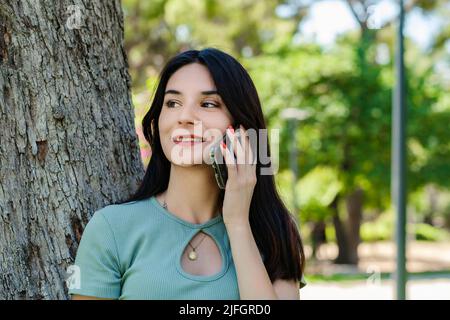 Bella donna bruna con t-shirt turchese in piedi sul parco della città, all'aperto parlando al telefono cellulare con gli amici o ragazzo con sorrisi. Foto Stock