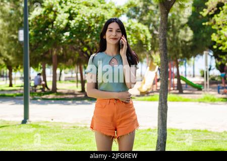 Donna bruna carina con t-shirt turchese in piedi sul parco della città, all'aperto parlando al telefono cellulare con amici o ragazzo con sorrisi. Foto Stock