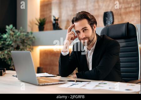Ritratto di un bell'attraente uomo d'affari caucasico positivo in una causa formale, ceo aziendale, top manager, seduto a una scrivania con un laptop in un ufficio moderno, guardando la fotocamera, sorridente Foto Stock