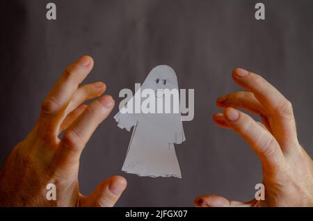 Mani di un uomo con le dita piegate intorno ad un fantasma di carta bianca. Attributo di Halloween tagliato da un foglio di carta con forbici. Volare in aria Ghost si Foto Stock