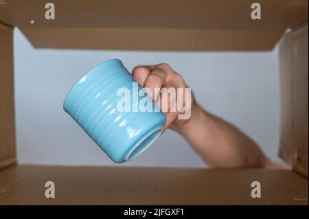 La mano di un uomo posiziona una tazza blu in una scatola mobile di cartone. Tazza in ceramica per bevande calde. Vista dal basso. Vista interna. Primo piano. Messa a fuoco selettiva. Foto Stock