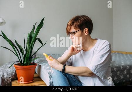 Donna con capelli corti a casa seduta sul pavimento con il suo smartphone Foto Stock
