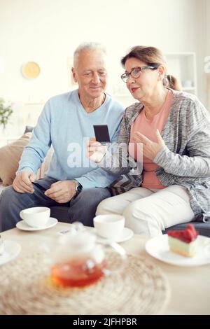Anziana signora attraente in occhiali seduti sul divano e gesturing mano mentre condividendo la storia del passato con il marito e mostrando vecchie fotografie a lui Foto Stock