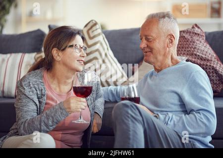 Felice bell'uomo dai capelli grigi in maglione appoggiato sul divano e bevendo vino mentre si fa una notte romantica con la moglie anziana Foto Stock