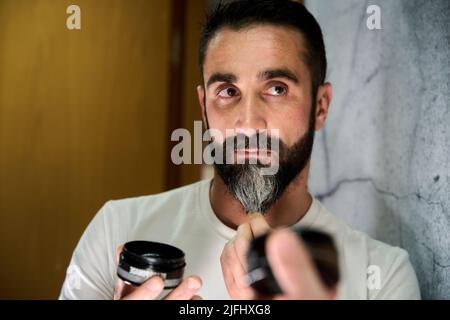 Giovane uomo che applica cera e olio alla barba davanti allo specchio. Foto di alta qualità Foto Stock