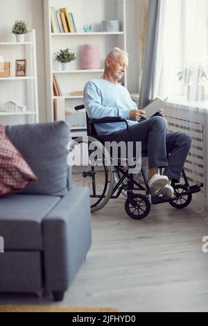 Uomo serio maturo dai capelli grigi con gambe paralizzate seduta in sedia a rotelle dalla finestra e libro di lettura Foto Stock