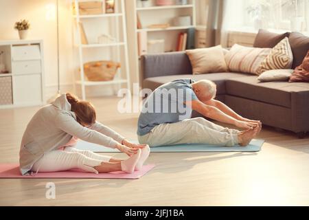 Coppia anziana in abbigliamento sportivo seduto su materassini da ginnastica in soggiorno e piegandosi in avanti mentre si allunga la schiena con yoga Foto Stock