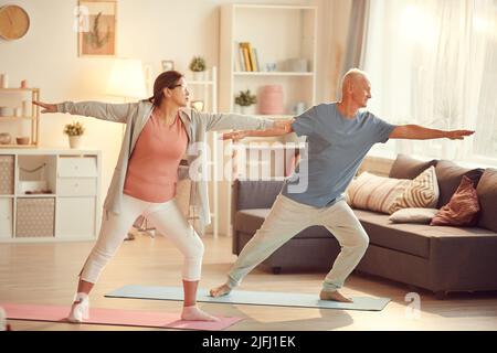 Coppia matura attiva in abbigliamento sportivo in piedi su materassini da ginnastica in soggiorno e braccia stretching mentre si pratica lo yoga insieme Foto Stock