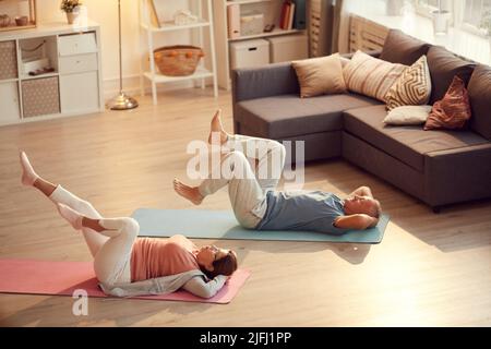 Coppia anziana sana sdraiata su tappetini in soggiorno e gambe rotanti durante l'esercizio per le articolazioni del ginocchio Foto Stock