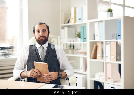 Ritratto di contenuto giovane successo bearded manager in gilet e cravatta seduta a tavola in ufficio e tenendo in mano planner con penna Foto Stock